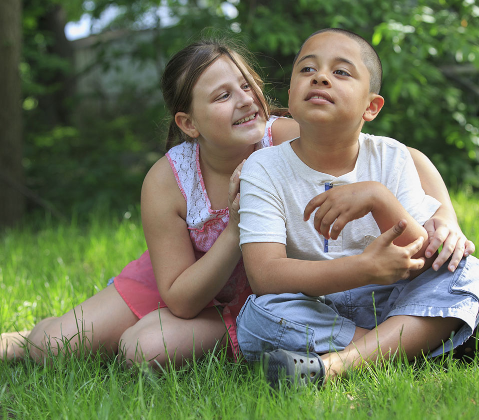Young smiling girl sits on grass with arm around boy who's looking off into the distance